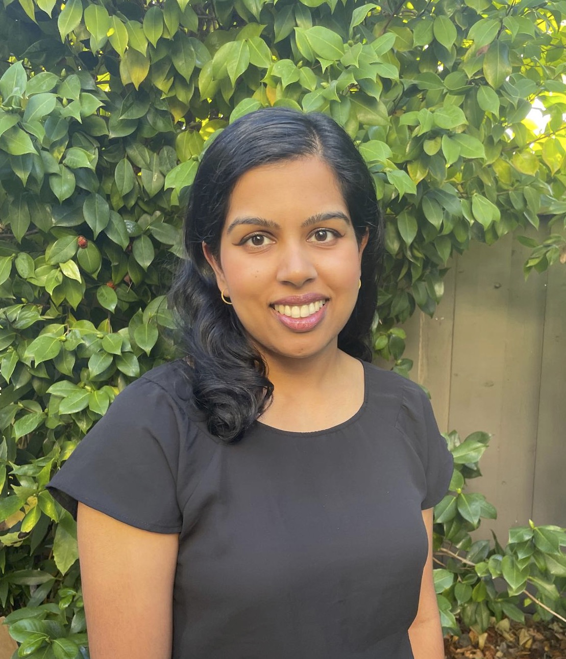 Headshot with Esha standing in front of some bushes. She is wearing a black t-shirt.