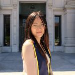 Avery standing outside of a building in a dress and graduation sash